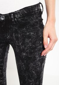 Černé džínové kalhoty s texturovaným povrchem, vybavené předním zapínáním na knoflíky, poutky na opasek a mírným vybledlým vzorem na látce.