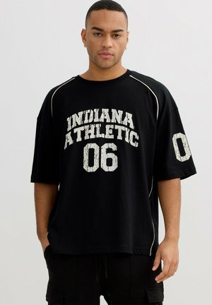 Hombre con el pelo corto que lleva una camiseta negra de gran tamaño con el texto blanco "INDIANA ATHLETIC 06" y el número 0 en la manga izquierda.