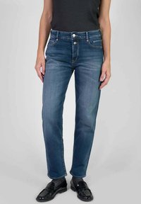 Persoon in een middelhoge, recht gesneden blauwe jeans, gecombineerd met zwarte instappers en een zwart mouwloos topje, staande tegen een effen achtergrond.