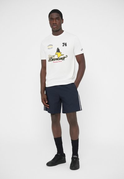 Άσπρο βαμβακερό T-shirt με γραφικό σχέδιο, που απεικονίζει έναν κιτρινο καρτούν χαρακτήρα και κείμενο. Συνδυάζεται με σκούρο μπλε σορτς και μαύρα αθλητικά παπούτσια.