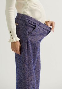 Blaue Hosen im Fischgrätenmuster mit weitem Bein, aus strukturiertem Stoff und tiefen Taschen, getragen von einer Person, die den Bund hält.