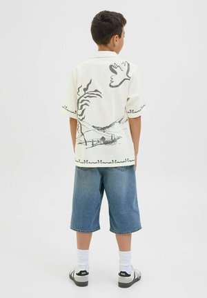 Chemise crème à manches courtes avec un design graphique noir, associée à un short en denim de longueur genou. Modèle portant des chaussettes blanches et des baskets noires.