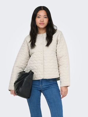 Jeune femme aux longs cheveux foncés portant une veste beige matelassée et un jean bleu, tenant un grand sac à main en cuir noir à ses côtés.