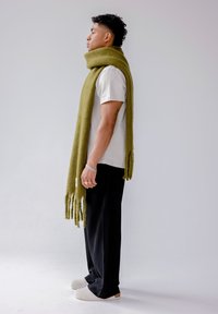 Écharpe vert olive avec détails à franges, mélange de laine, portée sur une chemise blanche, associée à un pantalon côtelé noir et des chaussures blanches à enfiler.