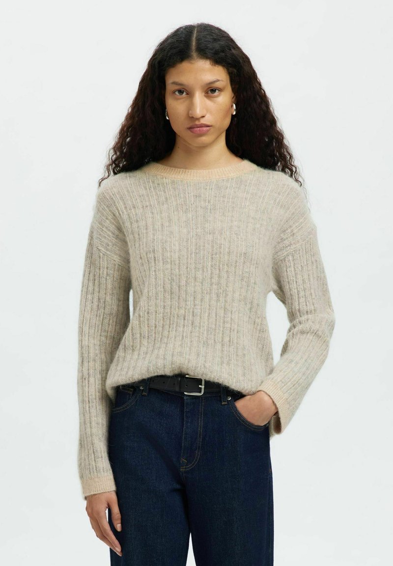 Femme avec de longs cheveux bouclés portant un pull côtelé beige rentré dans un jean bleu foncé avec une ceinture noire, se tenant devant un fond uni.