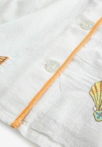 Balti lininiai marškiniai su oranžine apdaila, skaidriais mygtukais ir siuvinėtais jūros kriauklių bei vėduoklių motyvais. Audinys turi tekstūrą ir atrodo lengvas bei gaivus.