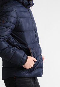 Chaqueta acolchada azul marino con cremallera frontal, capucha y bolsillos laterales. Presenta un diseño acolchado y acentos marrones en los bolsillos. Textura suave.