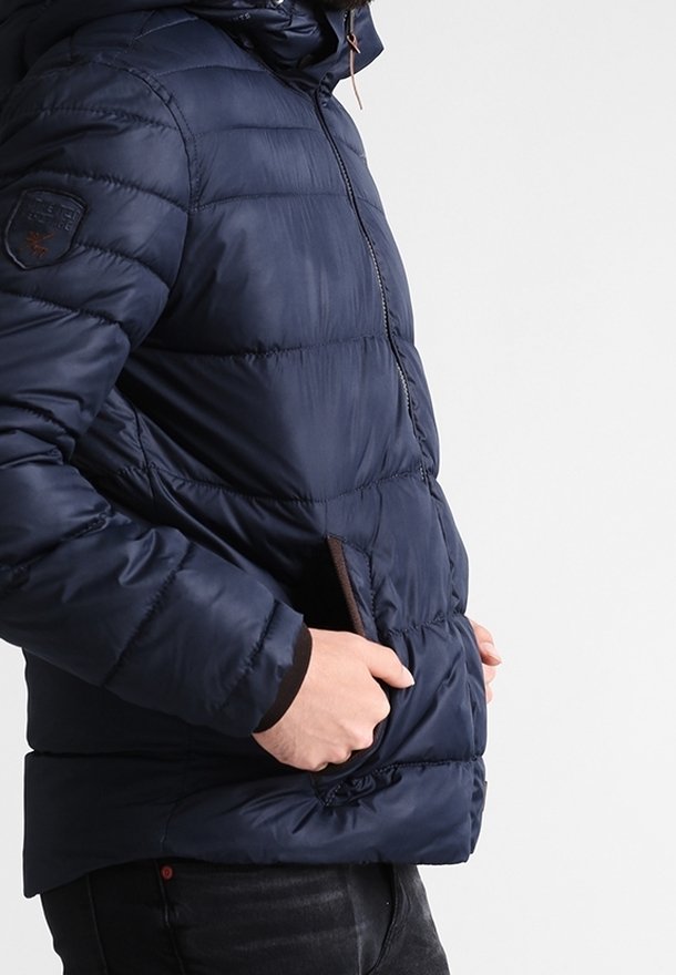 Chaqueta acolchada azul marino con cremallera frontal, capucha y bolsillos laterales. Presenta un diseño acolchado y acentos marrones en los bolsillos. Textura suave.