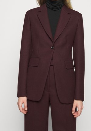 Vrouw draagt een donker bordeauxkleurig, op maat gemaakt blazer met bijpassende broek en een zwarte coltrui eronder, staand tegen een effen achtergrond.