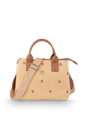 Woman in dark coat and beige sweater holding beige tote bag with small red flower patterns, standing among green plants outdoors.