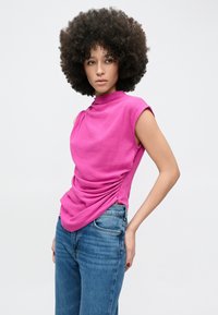 Top fucsia fără mâneci cu gât înalt și detaliu adunat pe lateral, asortat cu blugi din denim albastru deschis. Textură netedă.