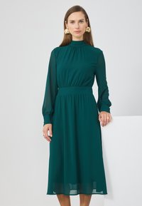 Robe longue à manches longues teal avec un col et une taille froncés, arborant une jupe fluide et des manches transparentes. Boucles d'oreilles circulaires dorées remarquables.