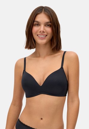 Mujer con cabello castaño hasta los hombros sonriendo, vistiendo un sujetador inalámbrico negro y ropa interior a juego contra un fondo claro y liso.