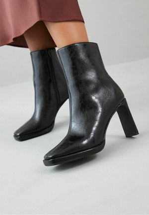 Bottines en cuir noir brillant à talons blocs portées avec une jupe marron, sur un fond de sol gris clair.