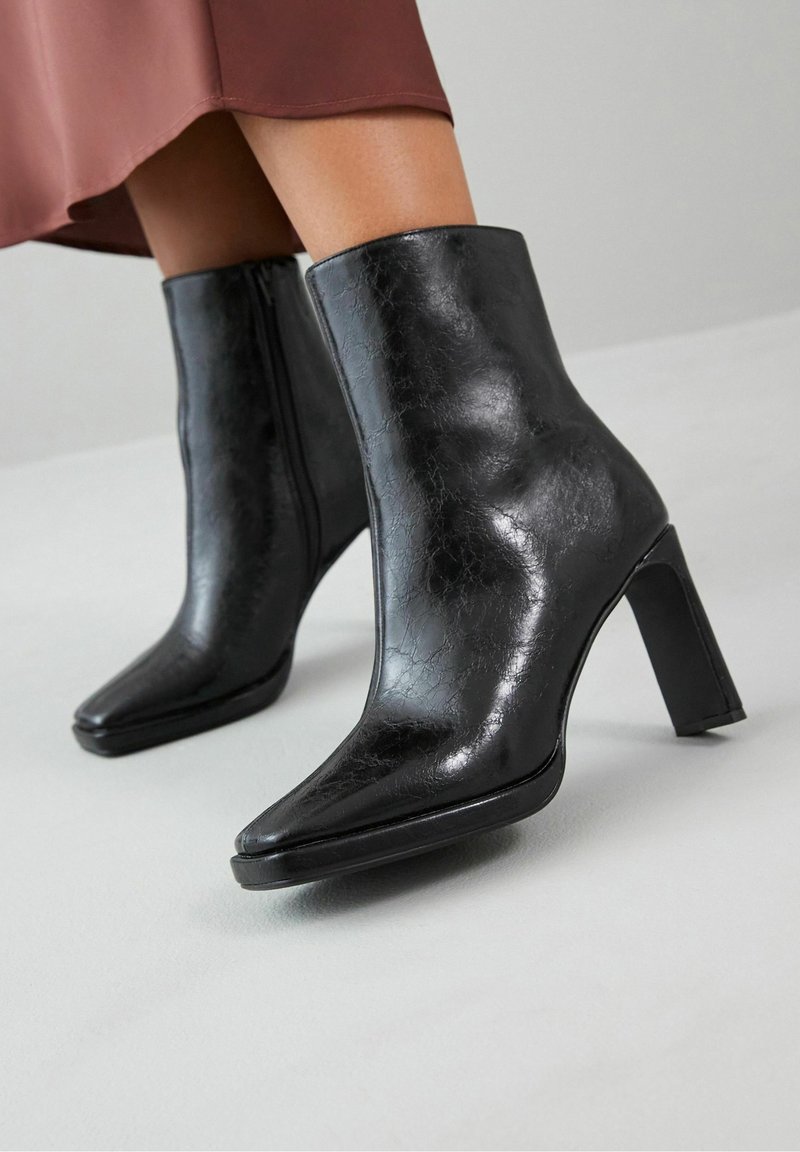 Bottines en cuir noir brillant à talons blocs portées avec une jupe marron, sur un fond de sol gris clair.