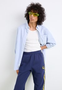 Camisa de algodão azul claro com riscas, por cima de uma t-shirt branca, combinada com calças de fato de treino azul-marinho com riscas amarelas e um emblema da Nike. Óculos de sol verdes.