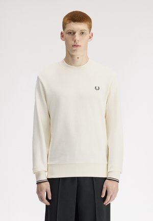 Junger Mann mit kurzem roten Haar trägt einen cremefarbenen Pullover mit einem kleinen schwarzen Lorbeer-Logo und schwarze plissierte Hose vor weißem Hintergrund.