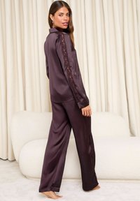 Completo pigiama di raso in un profondo viola con maniche lunghe, caratterizzato da dettagli in pizzo lungo le maniche e le cuciture laterali; vestibilità comoda con pantaloni dritti.