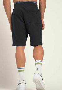 Mann in schwarzen Shorts mit Gesäßtasche, weißen Socken mit grünen und blauen Streifen und weißen Turnschuhen, der vor einem schlichten Hintergrund steht.