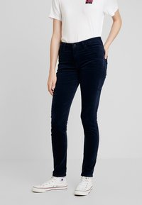 Marina kordflöjlsbyxor med en figursytt snitt, framfickor och en slät textur, kombinerade med en vit T-shirt och vita sneakers.