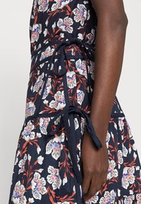 Vestido floral com fundo azul-marinho, flores brancas e vermelhas, detalhe de cintura amarrada, camadas de babados e textura suave de algodão.