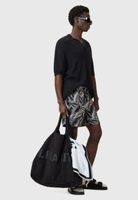 Polo shirt nero a maglia, pantaloni corti con motivo, occhiali da sole neri e una grande borsa nera con un asciugamano bianco. Sandali con fibbie.