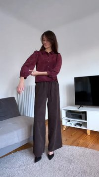 Burgundy button-up blouse with cuffed sleeves, paired with brown wide-leg trousers. Model wears black pointed heels in a minimal indoor setting.