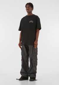 Sort oversized bomulds t-shirt med logo og korte ærmer, parret med løstsiddende mørkegrå cargobukser med flere lommer. Sorte sneakers.