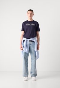 Marineblauwe katoenen t-shirt met "Calvin Klein" logo, lichtblauw shirt geknoopt om de taille, lichtgewassen wijdere broek en witte sneakers.