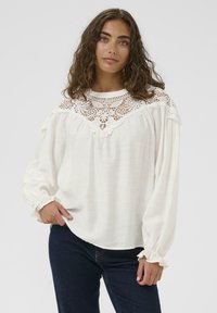 Blusa bianca con dettagli in pizzo attorno al collo. Maniche lunghe con polsini arricciati, realizzata in un tessuto leggero e testurizzato.