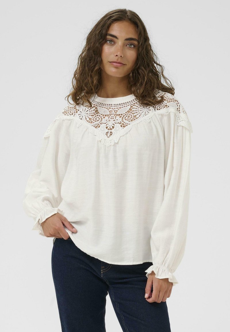 Blusa bianca con dettagli in pizzo attorno al collo. Maniche lunghe con polsini arricciati, realizzata in un tessuto leggero e testurizzato.