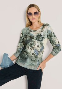Vrouw draagt ronde zonnebril, bloemmotief lange mouwen top in groentinten, donkere gestructureerde broek en houdt een lichtblauwe clutch tas vast.