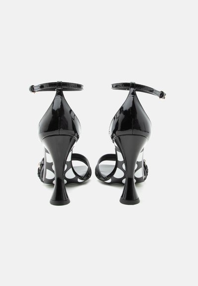 Ζευγάρι μαύρες λουστρίνι ψηλοτάκουνες σανδάλες με λουράκια στον αστράγαλο και λευκά πουά στην σόλα, φαίνονται από πίσω.