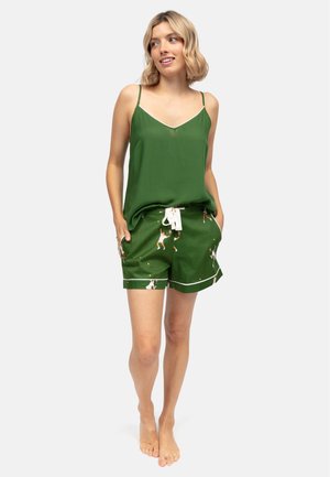 Vrouw draagt een groene top en groene shorts met een koord, bedrukt met kleine witte en oranje honden, staat blootsvoets met handen in haar zakken.