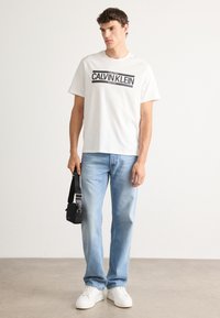 Young man wearing white Calvin Klein logo t-shirt, light blue jeans, white sneakers, holding small black shoulder bag by side.