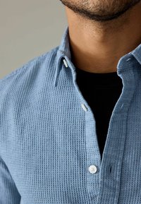 Cămașă texturată albastră cu guler tip button-down, având nasturi albi și un model subtil în formă de wașă, stratificată peste o cămașă neagră.