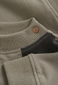 Olivgrüner Pullover mit ribbeltem Kragen und kontrastierendem schwarzen Einsatz. Kupferne Knopfdetails sorgen für einen dezenten Akzent. Weicher Baumwollstoff.