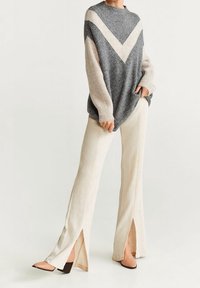 Pull en tricot gris et beige avec un motif en V, associé à un pantalon évasé beige avec des fentes sur les côtés et des chaussures à talons carrés marron.