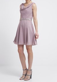 Fialová bezramínková šaty s drapovaným výstřihem, přiléhavým živůtkem a rozšířenou sukní. Disponuje stříbrným zdobeným páskem a třpytivými sandály na kotníkovém pásku.
