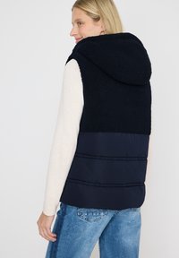 Chaleco azul marino con capucha, que presenta una parte superior de textura esponjosa y un fondo de material acolchado suave. Diseño sin mangas, con costuras para definir la forma.