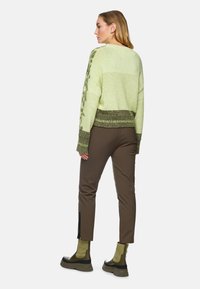 Grüner Strickpullover mit einem strukturierten Muster, kombiniert mit braunen Slim-Fit-Hosen und grünen Ankle-Boots mit dickem Sohlen und seitlichen Reißverschlüssen.