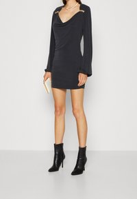 Abito nero a collo alto con maniche lunghe, dettagli in bottoni dorati sulle spalle e vestibilità aderente. La modella abbina stivali neri alla caviglia e una clutch.