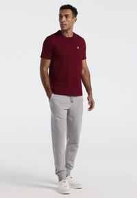 Bordeaux-rotes T-Shirt mit kurzen Ärmeln und Brusttasche, kombiniert mit hellgrauen Sweatpants mit Kordelzug und elastischen Bündchen. Weiße Sneakers.