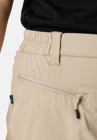 Beige lightweight shorts with an elastic waistband, featuring a zippered pocket and a small blue and yellow label on the side.