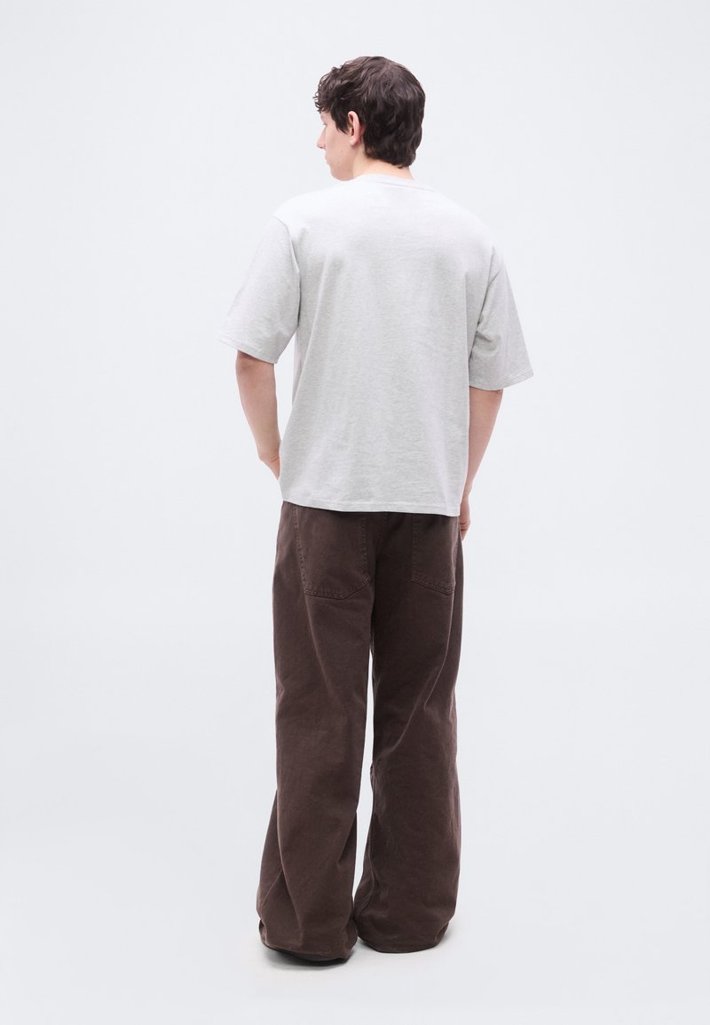 Giovane uomo visto di spalle, indossa una T-shirt oversize grigio chiaro e pantaloni marroni a gamba larga su sfondo bianco.