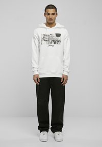 Weißer Hoodie mit einem grafischen Druck von Händen und dem Wort "pray" in Schwarz in der Mitte. Lockere Passform, kombiniert mit schwarzen Hosen und weißen Sneakers.