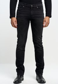 Jeans in denim neri con vestibilità slim, dotati di cinque tasche, gambe dritte e una texture liscia. Indossati con stivali neri alla caviglia.