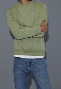 Felpa verde chiaro con collo rotondo e polsini a coste, caratterizzata da un piccolo logo "blend" sul petto. Indossata con jeans blu.