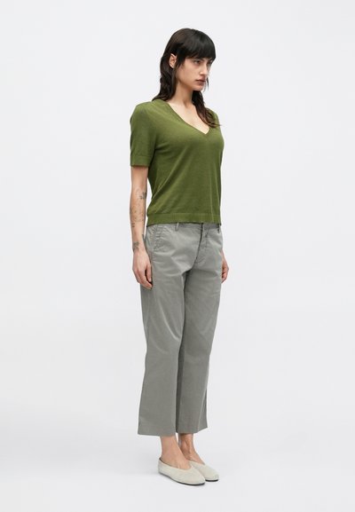 Πράσινο πουλόβερ με κοντά μανίκια σε σχήμα V και άνετη εφαρμογή, συνδυασμένο με γκρι παντελόνι με φαρδιά μπατζάκια και ανοιχτόχρωμα παπούτσια slip-on.
