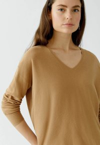V-Ausschnitt, langärmliges Oberteil aus strukturierter, gerippter Baumwolle in einem warmen, camel Farbstich. Lässige Passform mit umgeschlagenen Ärmeln und seitlichen Taschen.